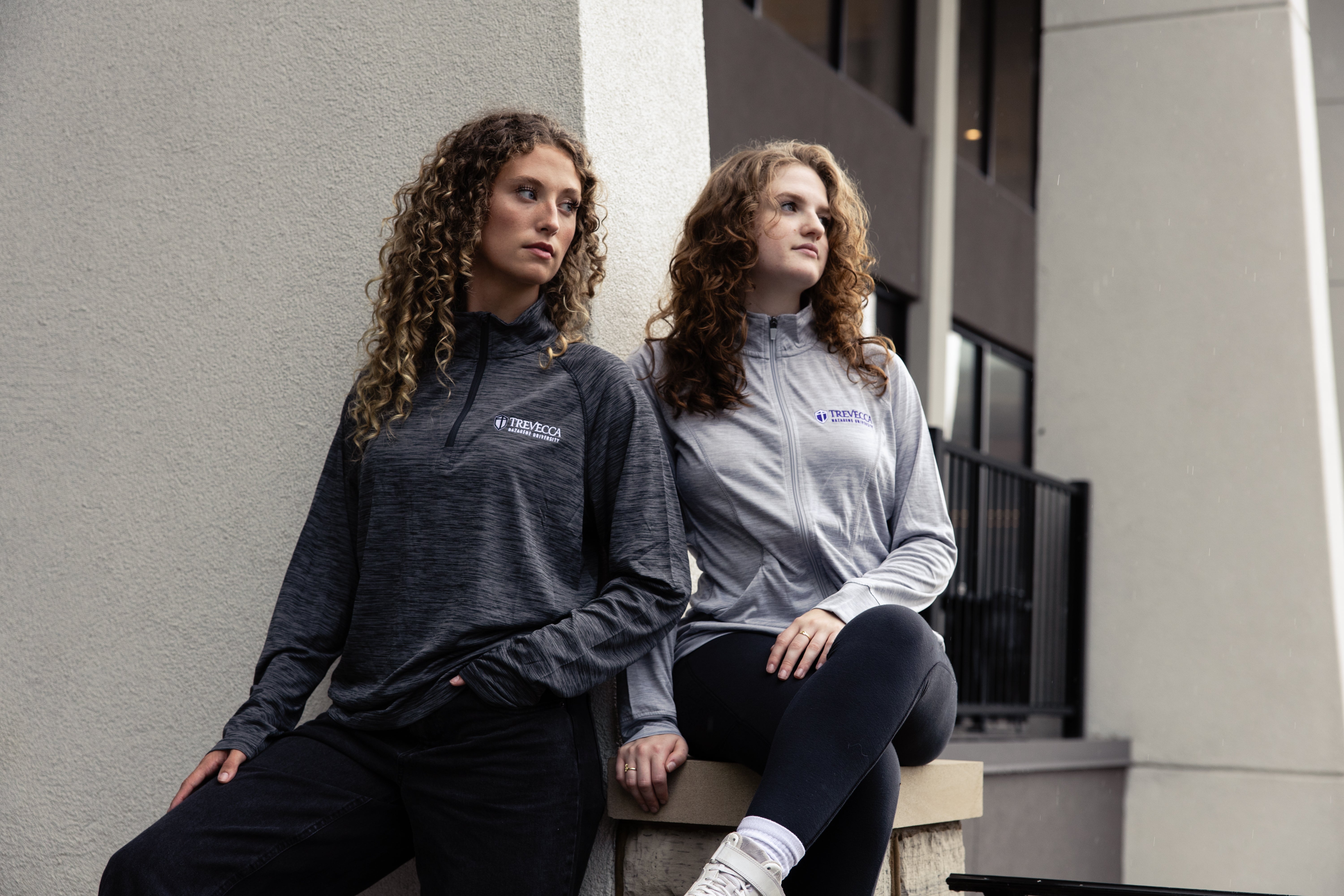 Two women wearing gray athletic jackets with Trevecca logos, sitting on a ledge outdoors.